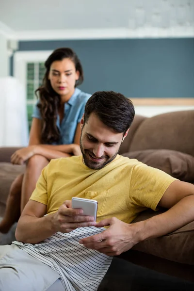 Mujer celosa mirando al hombre usando el teléfono móvil en la sala de estar —  Fotos de Stock