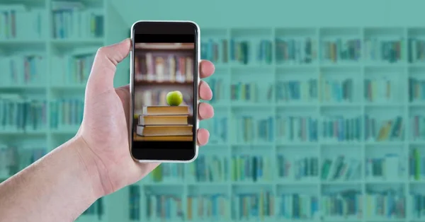 Hand with phone showing book pile with apple against blurry bookshelf with blue overlay — Stock Photo, Image
