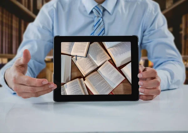Business man at table with tablet showing open books against blurry bookshelves — Stock Photo, Image