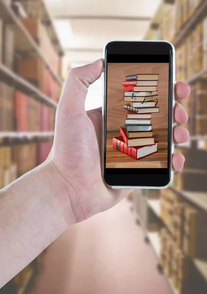 Hand with phone showing book pile against blurry bookshelfs — Stock Photo, Image