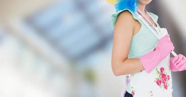 Woman in apron with duster against blurry window — Stock Photo, Image