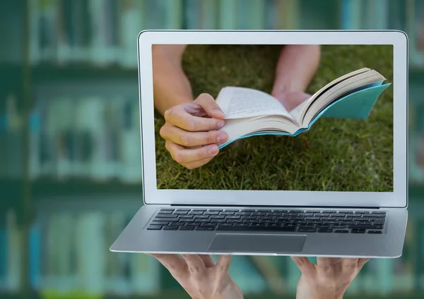 Hands with laptop showing book on grass against blurry bookshelf with green overlay — Stock Photo, Image