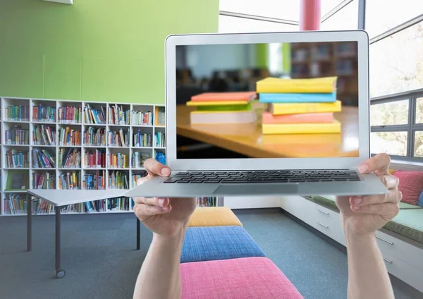 Mani con computer portatile che mostra pile di libri contro la biblioteca — Foto Stock