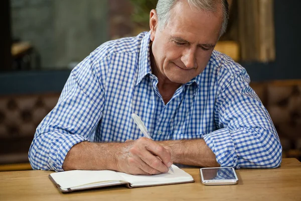 Starší muž psaní na deník u stolu — Stock fotografie