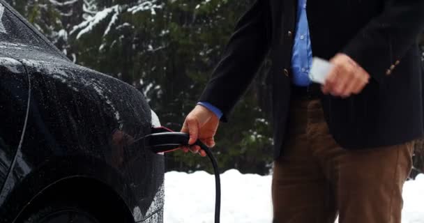 Чоловік використовує мобільний телефон і заряджає електромобіль у сніжний день — стокове відео