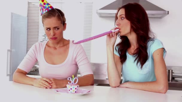 Woman blowing party horns beside her upset friend in kitchen — Stock Video