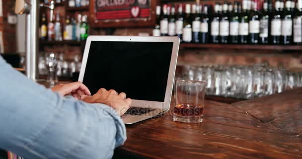 Человек, использующий ноутбук, выпивая виски за стойкой — стоковое видео