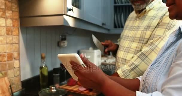 Starszy człowiek do krojenia warzyw, podczas gdy kobieta za pomocą cyfrowego tabletu w kuchni — Wideo stockowe