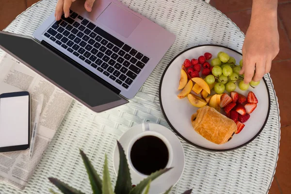 Manos recortadas que tienen fruta mientras se utiliza el ordenador portátil —  Fotos de Stock