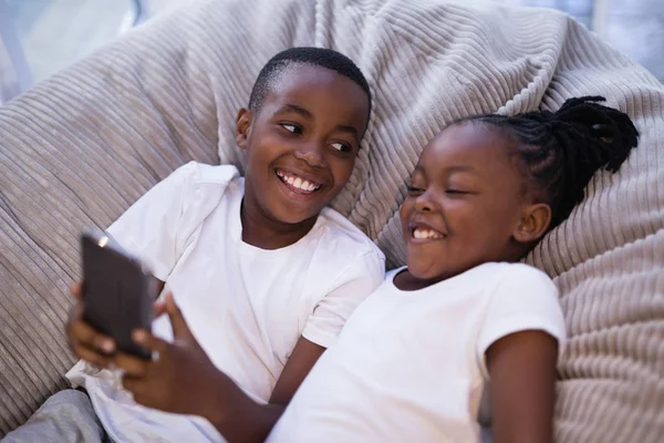 Hermanos usando el teléfono mientras están acostados en el sofá — Foto de Stock