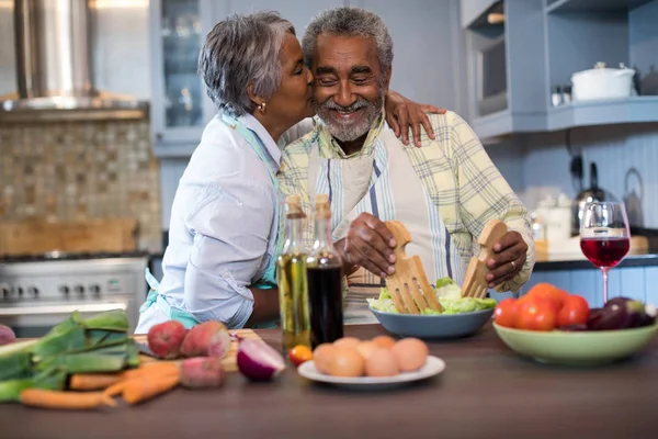 Senior mulher beijando o homem enquanto prepara a comida — Fotografia de Stock