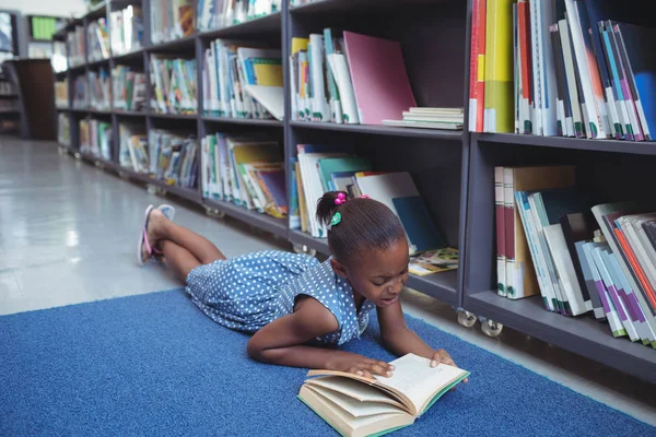 Menina lendo livro por prateleira na biblioteca — Fotografia de Stock