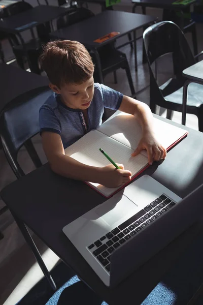 Junge schreibt in der Schule mit Laptop in Buch — Stockfoto