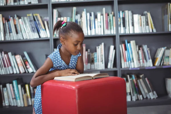 Girl reading book on ottoman in library — Stock Photo, Image