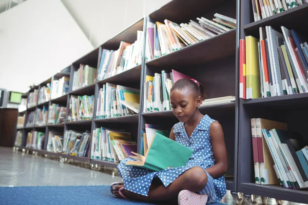 Menina lendo livro por prateleira na biblioteca — Fotografia de Stock