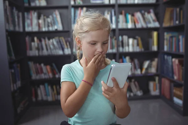 Überraschtes Mädchen schaut in Bibliothek auf Smartphone — Stockfoto