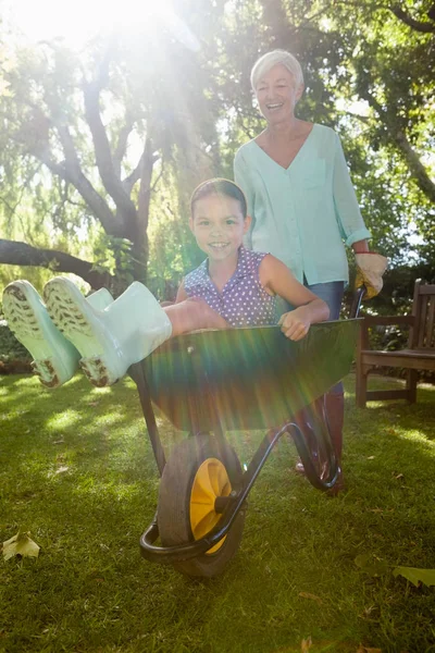 Smiling grandmother pushing granddaughter in wheelbarrow — Stock Photo, Image