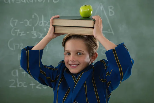 Colegial sosteniendo libros pila con manzana en la cabeza contra pizarra —  Fotos de Stock