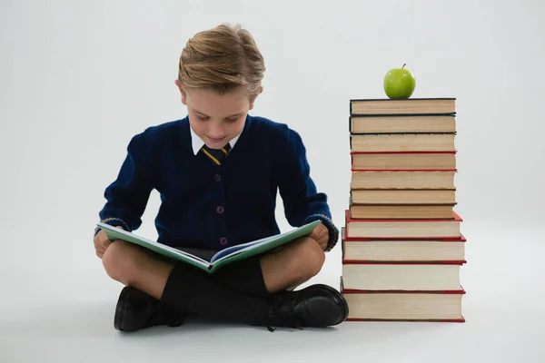 Schoolboy reading book while sitting beside books stack — Stock Photo, Image