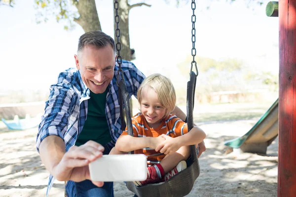 Padre tomando selfie con hijo sentado en swing — Foto de Stock