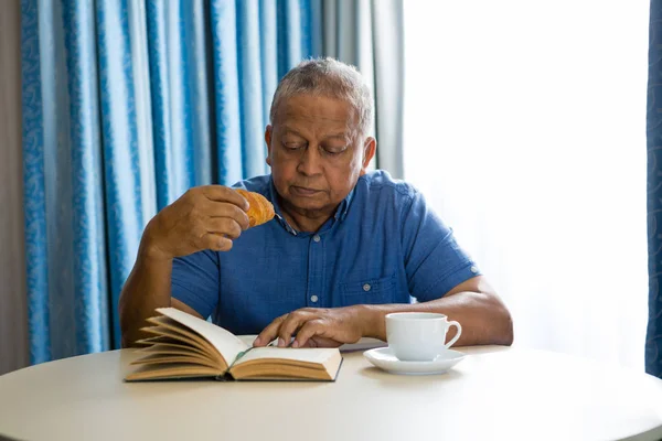 Homem sênior comendo croissant enquanto lê livro — Fotografia de Stock