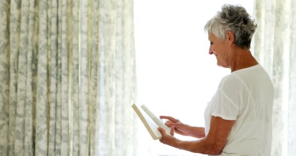 Улыбающаяся пожилая женщина читает книгу в спальне — стоковое видео
