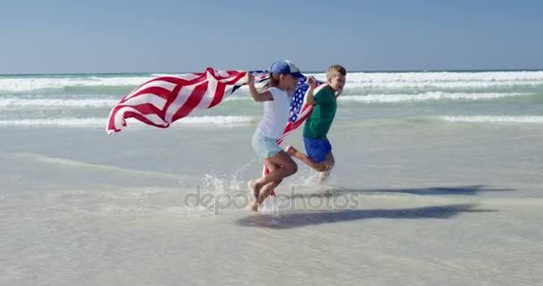 Siblings holding American flag while running on shore at beach — Stock Video