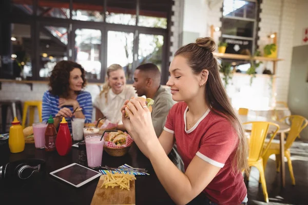 Mujer comiendo hamburguesa mientras está sentado en la cafetería —  Fotos de Stock
