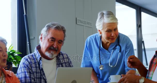 Enfermera sirviendo taza de té mientras amigos mayores utilizando el ordenador portátil — Vídeo de stock