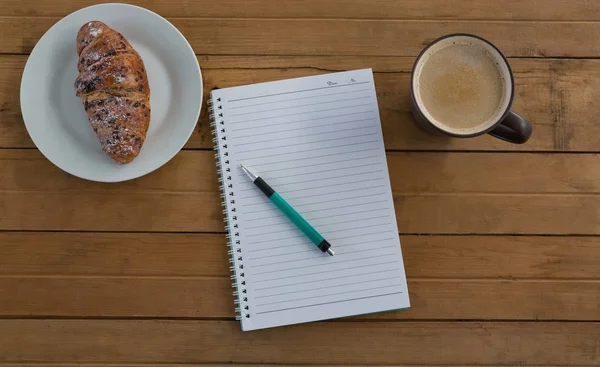 Croissant, café, bolígrafo, bloc de notas sobre tabla de madera — Foto de Stock