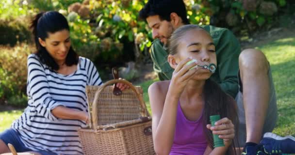 Flicka Som Blåser Bubble Medan Hennes Föräldrar Som Sitter Bakgrunden — Stockvideo