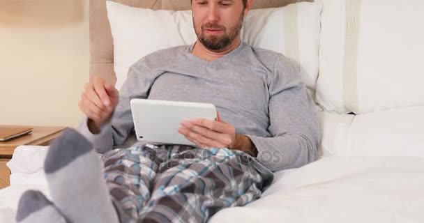 微笑的年轻人坐在床上使用他的平板电脑在家里的卧室4K — 图库视频影像