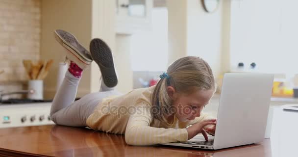 のキッチンで彼女のラップトップを使用してテーブルの上に横たわるの幸せな女の子 — ストック動画