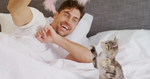の寝室で彼のトラ猫で遊んでベッドで横になっている若い男の笑みを浮かべてください — ストック動画