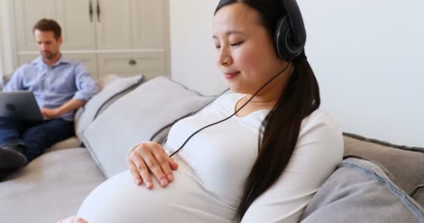 を自宅のリビング ルームでヘッドフォンで音楽を聞いて 妊娠中の女性 — ストック動画