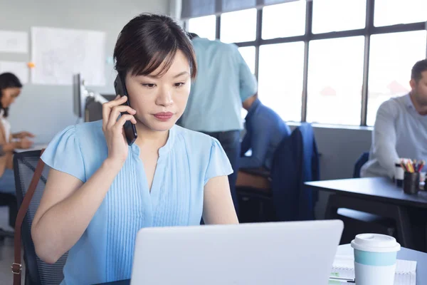 アジアの女性ビジネスの最前線カジュアルモダンなオフィスで働くクリエイティブな仕事をしている女性が ノートパソコンを使ってデスクに座り スマートフォンで話をしています — ストック写真