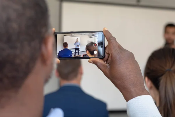 回顾一位非裔美国男性商业创意者手持智能手机 在商业会议上与其他听众一起拍摄演讲者的情景 — 图库照片