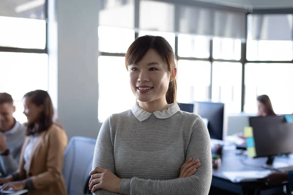 在一个随意的现代办公室里工作的一位亚洲女企业家的画像 她抱着胳膊站在那里 微笑着 把目光投向别处 身后是同事们的作品 — 图库照片