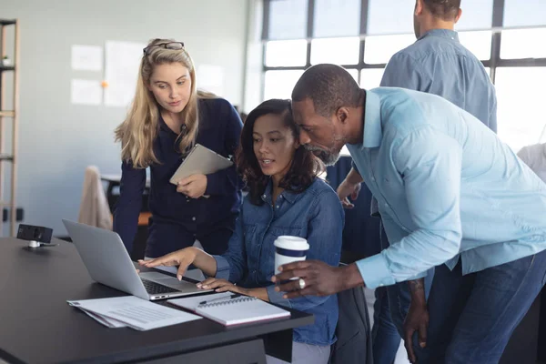 Side view of an African American male and a Caucasian female business creative standing either side of a mixed race female colleague sitting a desk using a laptop and discussing her work with them, in a casual modern office