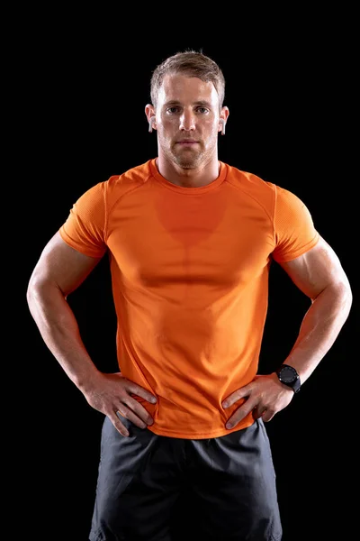 一名身穿运动服 头戴无绳耳机 体格健壮的白人男子在锻炼后站在前视镜前 双手放在臀部上 黑色背景上的垂直射击 — 图库照片