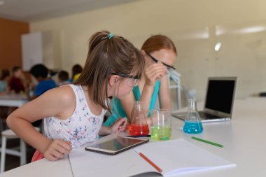 Side view of two Caucasian schoolgirls sitting at a desk wearing safety glasses and using flasks of colourful liquid while doing an experiment during an elementary school science class, with classmates working together at a table in the background clipart