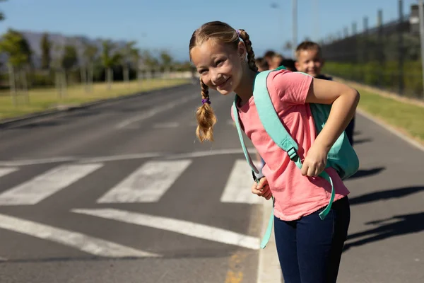 ピンクのTシャツを着てリュックサックを担い 晴れた日に小学校への道を横断するのを待っている間に交通を探している白人の女の子のフロントビューを閉じます — ストック写真