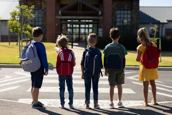 在阳光明媚的日子里 一群三个白人学童和一个拿着背包的女学生在过马路的人行横道上等待着他们小学门前的后视镜 — 图库照片