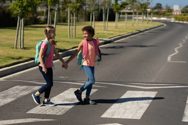 Side view of a Caucasian and an African American schoolgirls wearing rucksacks holding hands and crossing the road at a pedestrian crossing on their way to elementary school on a sunny day