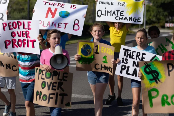 Front view of a diverse group of elementary school pupils on a protest march, carrying signs with environmental and conservation slogans on them, one girl shouting in a megaphone as they walk down a road in the sun