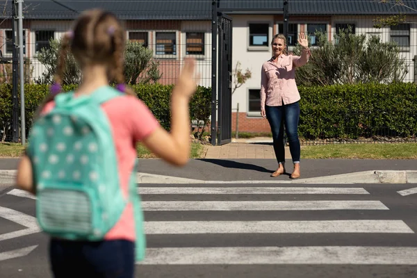 晴れた日には小学校への道を横断する歩行者の反対側から母親に手を振ってバックパックを身に着けている白人の女子学生の背面図 — ストック写真