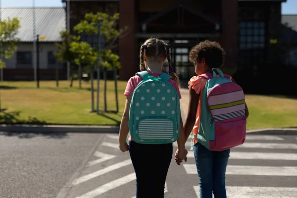 阳光灿烂的一天 一个白人和一个非洲裔美国女学生手牵着背包 在去小学的路上穿过马路的后视镜 — 图库照片