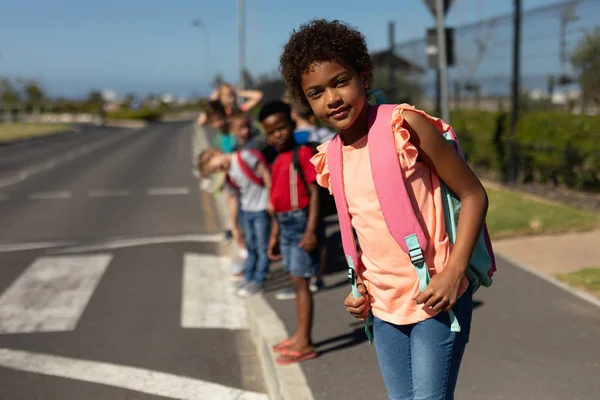 ピンクのTシャツを着てリュックサックを担い 晴れた日に小学校への道を横断するのを待っている間に交通を探しているアフリカ系アメリカ人の女子学生のフロントビュー — ストック写真