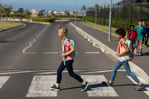 Side view of a Caucasian and an African American schoolgirl wearing rucksacks crossing the road at a pedestrian crossing on their way to elementary school on a sunny day, with other schoolchildren in the background