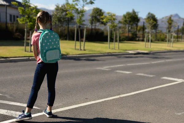 在阳光明媚的日子里 一个头戴斑斑头戴背包的白人女学生在过马路的人行横道上等着上小学的后视镜 — 图库照片